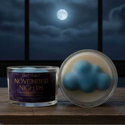 A candle product shown in a jar with a dark  label reading 'November Nights,' alongside an open round lid revealing a blue cloud-shaped wax piece on a cream base, set on a wooden surface against a night-sky scene with the moon.